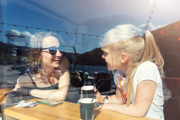 Through window view of young adult happy mom enjoy having fun talking, drink hot chocolate laugh with cute little daughter sitting at cafe restaurant on sunny day. Mother and child friendship concept