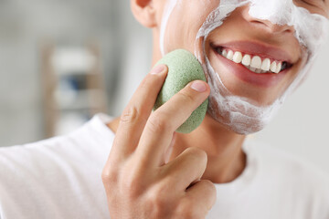 Poster - Happy young man washing off face mask with sponge in bathroom, closeup