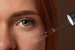 Beautiful young woman applying cosmetic serum onto her face on brown background, closeup