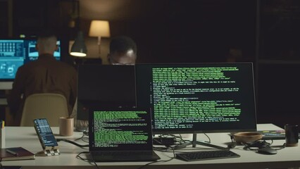 Lines of html code on computer and laptop screens at table in dark office with two male programmers sitting at their workplaces and developing new software
