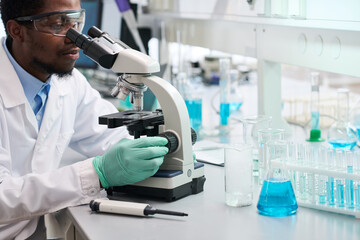 Side view at African American male scientist in safety goggles at laboratory desk with medical glassware studying samples with microscope