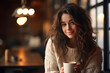 portrait of a young happy woman in a knitted sweater drinking coffee in a cozy cafe interior with lights bokeh