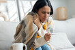 © nenetus - Tired woman with a cold taking her fever while lying with a blanket on the couch at home