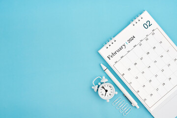 February 2024, Monthly desk calendar for 2024 year and alarm clock with paper clips on blue color background.