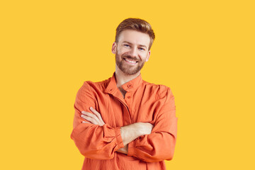 Wall Mural - Portrait of confident smiling young bearded handsome man standing with crossed arms wearing casual bright orange summer shirt isolated on studio yellow background. Smart guy looking at the camera.