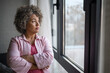 © lordn - A mature woman of mixed race looking outside a large window, her expression reflecting sadness, concern, and a sense of depression