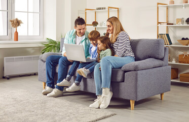 Happy young family with two kids girl and boy sitting on sofa using modern laptop together. Smiling parents resting on couch enjoying weekend watching funny cartoon online or talking on video call.