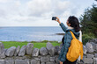 Latin woman traveler with a yellow backpack next to a gray stone wall taking a photo in a beautiful landscape with the sea in the background and the green field in ireland