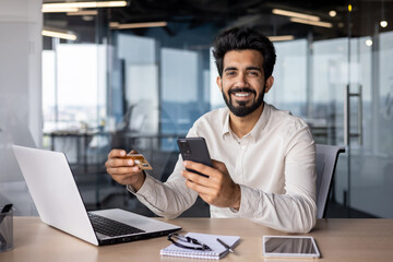 Wall Mural - Portrait of a smiling young man working in the office, sitting at a desk with a laptop, holding a credit card and a phone, smiling and looking at the camera