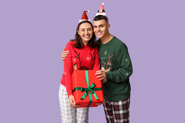 Wall Mural - Happy young couple in Christmas pajamas and with gift on lilac background