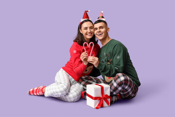 Wall Mural - Happy young couple in Christmas pajamas, with gift and candy canes on lilac background