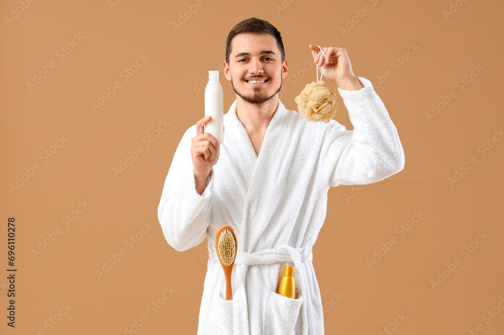Handsome young man in bathrobe with bath sponge and shower gel on beige background