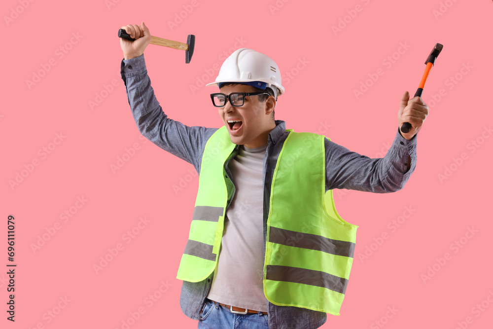 Angry male worker in eyeglasses with hammers on pink background