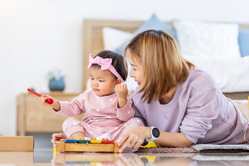 Asian mother is playing with her pretty smiling baby daughter with wooden toy block while spending quality time in the bed for family happiness and parenting