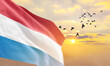 Waving flag of Luxembourg against the background of a sunset or sunrise. Luxembourg flag for Independence Day. The symbol of the state on wavy fabric.