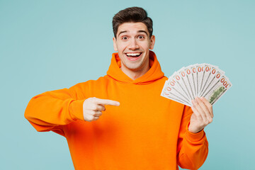 Poster - Young man he wears orange hoody casual clothes hold in hand point finger on fan of cash money in dollar banknotes isolated on plain pastel light blue cyan color background studio. Lifestyle concept.