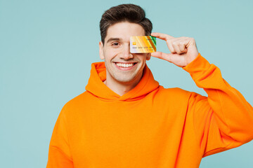 Poster - Young fun man he wears orange hoody casual clothes hold in hand cover eye with mock up of credit bank card isolated on plain pastel light blue cyan color background studio portrait. Lifestyle concept.