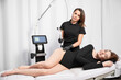 Cosmetologist using radiofrequency microneedling device while performing skin tightening treatment on female hip. Woman lying near cosmetology equipment and having stretch marks removal procedure.