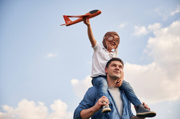 Sitting on shoulders, playing with toy plane. Father and little son are having fun outdoors