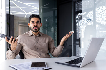 Portrait of a young angry and disappointed Indian man sitting at an office desk, holding a mobile phone and spreading his hands to the camera.