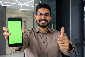 Close-up portrait of a young Indian man wearing glasses sitting in the office, showing the super sign with his finger to the camera and a green phone screen mockup.