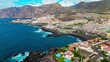 Aerial view of tourists enjoying Playa de la Arena a popular cove with a wide stretch of black sand. Luxury resort and colourful hotels with beautiful beaches in Puerto de Santiago, Tenerife.
