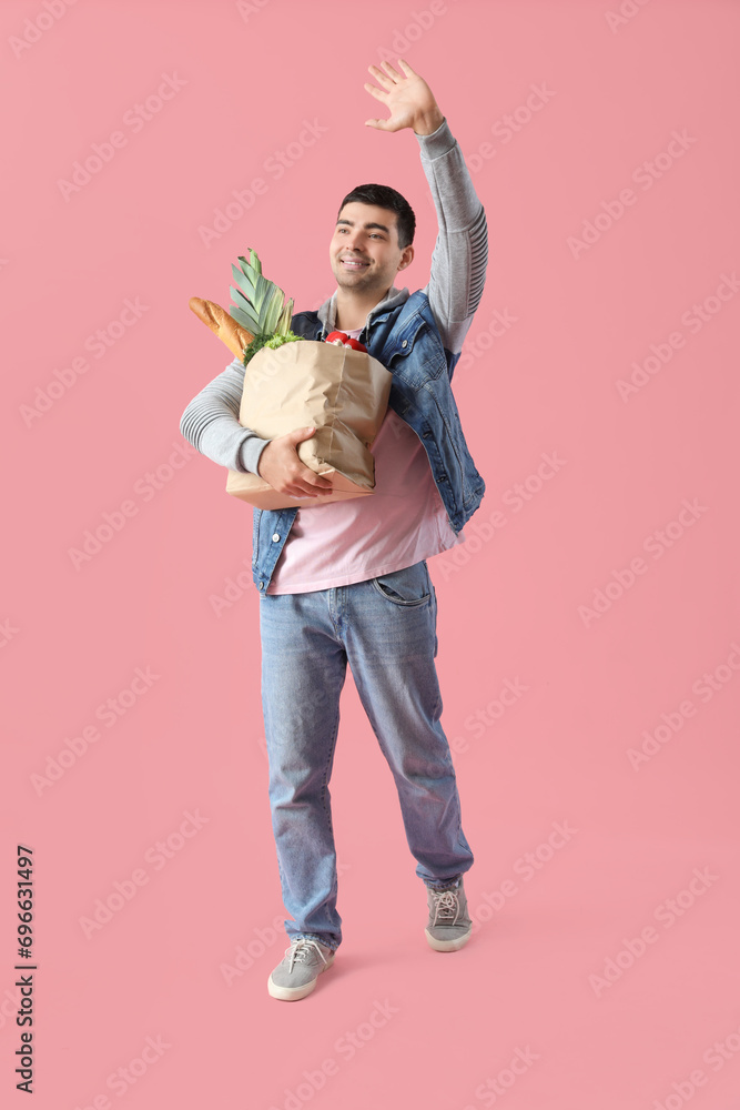 Young man with grocery bag waving hand on pink background
