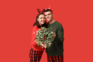 Wall Mural - Happy young couple in pajamas and with Christmas wreath on red background
