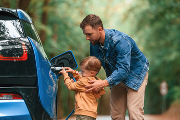 Teaching the boy where to put the wire. Father with his little son are waiting for electric car to charge