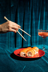 Background of hand holding sushi roll with chopsticks on red plate on dark blue background. Food composition.