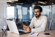 © Tetiana - Serious Muslim young man working in office, sitting at desk and using phone, texting, checking mail and bills, making phone calls