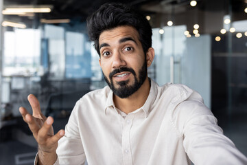 Wall Mural - Close-up photo of a young Indian man working in the office and talking on a video call, gesturing with his hand, holding a camera