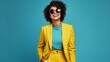 Full body happy cheerful Afro American woman wearing stylish yellow suit and trendy glasses standing isolated on blue background, looking at camera, winking her eye and smiling