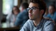 A young man in glasses thoughtfully listens during a meeting at a busy cafe with peers in the background.