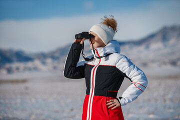 Wall Mural - Young woman tourist looking through binoculars on mountains background in winter day