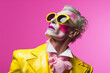 Older aged model, eccentric man in a vibrant costume, wearing makeup, trendy yellow sunglasses, suit and bow tie, on pink background. Concept of strange weird clown, irrational and weird behavior. 