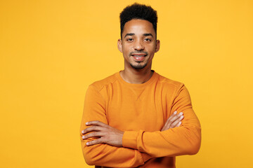 Wall Mural - Young smiling fun man of African American ethnicity wear orange sweatshirt casual clothes hold hands crossed folded look camera isolated on plain yellow background studio portrait. Lifestyle concept.