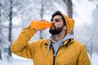 Exhausted handsome sportsman wearing yellow beanie and jacket drinking water from bottle after jogging in winter season.