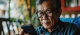 Fototapeta  - Elderly Asian man with eyesight issues removes glasses while using smartphone after browsing internet or social media at home.