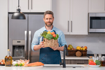 Wall Mural - Portrait of chef man in cook apron with a plate of vegetable salad in the kitchen. Man cooking vegan healthy salad in kitchen. Millennial man at modern kitchen with vegetables, prepare fresh vegetable