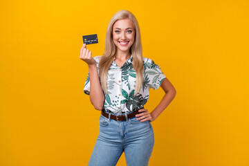 Sticker - Photo of positive girl with long hair dressed print shirt hand on waist demonstrate credit card isolated on yellow color background