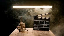 A Clapper Board And Vintage Video Camera On A Wooden Table Enveloped In Smoke. Cinematography Equipment In A Studio On A Black Background Illuminated By A Neon Lamp.