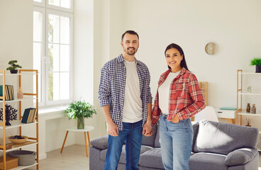 Wall Mural - Portrait of happy loving family couple at home. Young man and woman in jeans and plaid and checkered shirts standing together and holding hands in modern living room interior with couch in background