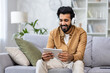 © Liubomir - Indian young man sitting on the couch at home wearing headphones and using a tablet with a smile.