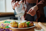 Fototapeta  - Hands of young unrecognizable woman sieving sugar powder on top of appetizing homemade donuts on plate while preparing for Mardi Gras