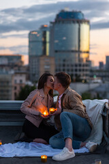 Wall Mural - Beautiful young loving couple on a surprise romantic date on a roof top. Picturesque view, skyscrapers on background. Wine, candles, kisses on holiday Saint Valentine's Day celebration outdoors