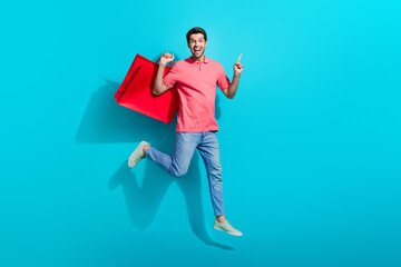 Sticker - Full length photo of funky cheerful man wear pink t-shirt jumping holding shoppers pointing empty space isolated blue color background