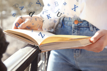 Wall Mural - Woman reading book with letters flying over it outdoors on sunny day, closeup