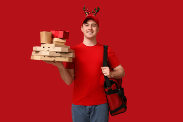 Sticker - Portrait of young delivery man in Christmas reindeer headband with bag, pizza boxes and present on red background