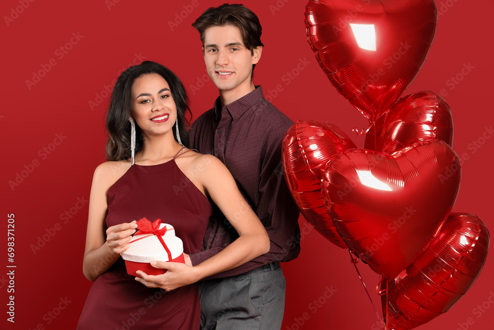 Young couple with gift box and balloons on red background. Valentine's Day celebration